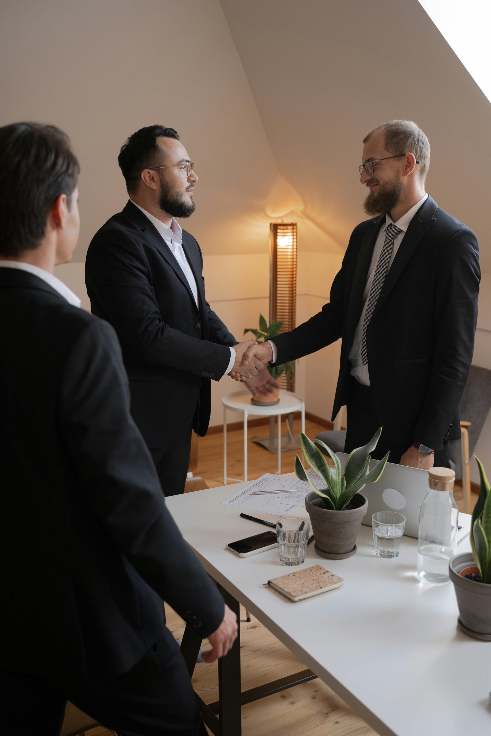 Home Businessmen in formal attire shaking hands during a meeting in a modern office setting.