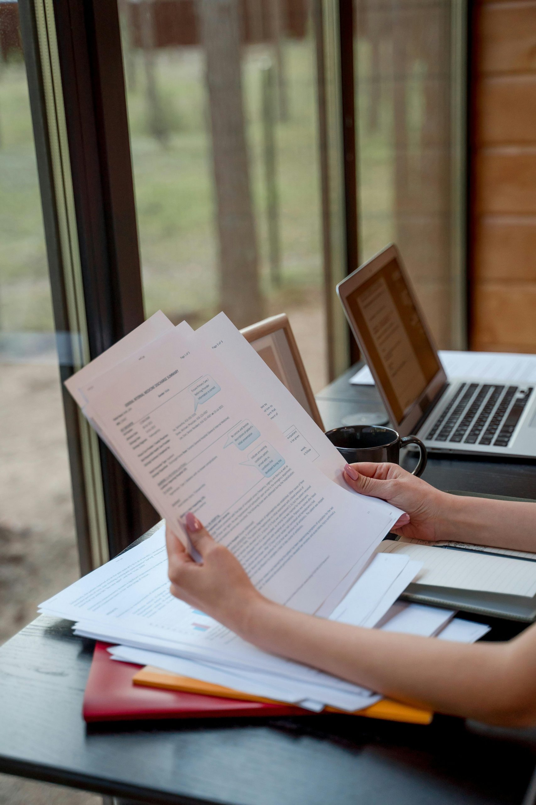 Person holding and reviewing documents near window with laptops on a desk.